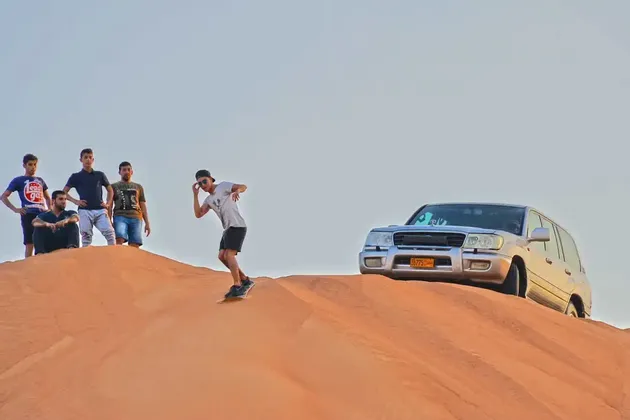 Ein WeRoad-Reisender sandboardet eine riesige Wüstendüne hinab, während seine Freunde und ein silberner SUV von oben zusehen.