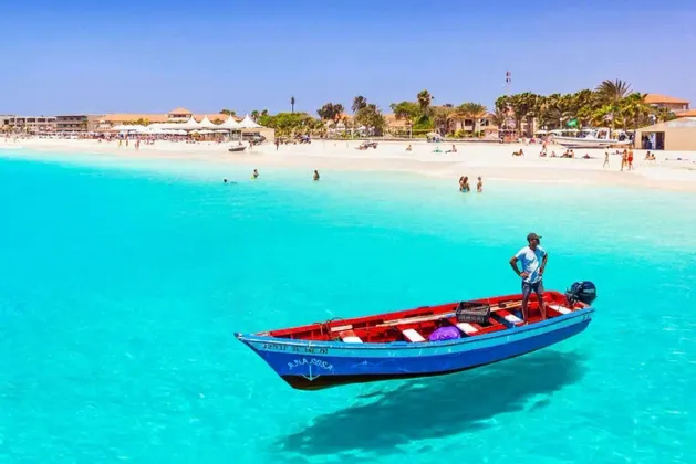 Un uomo è in piedi su una piccola barca blu che sembra galleggiare sull'acqua limpida e turchese vicino a una spiaggia sabbiosa.