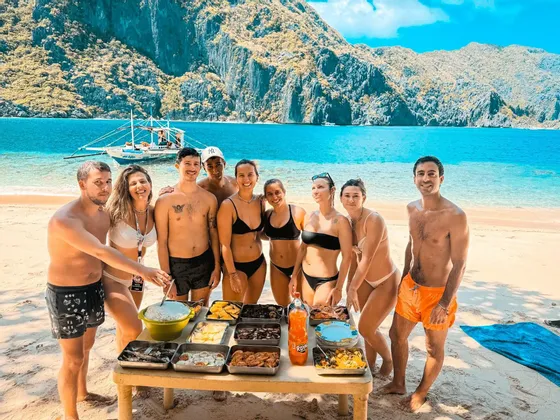 Un gruppo WeRoad in viaggio si raduna attorno a un tavolo imbandito su una spiaggia sabbiosa, con scogliere rocciose e una barca sullo sfondo.