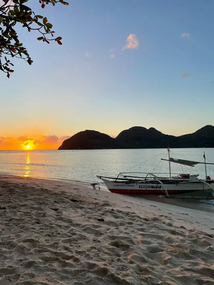 Una barca a bilanciere bianca si trova su una spiaggia sabbiosa mentre il sole tramonta sull'oceano, con colline in silhouette sullo sfondo.