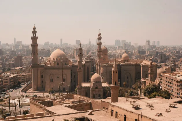 Une vue aérienne d'une grande mosquée couleur sable avec plusieurs dômes et minarets sur fond de paysage urbain brumeux avec des gratte-ciel modernes en arrière-plan.