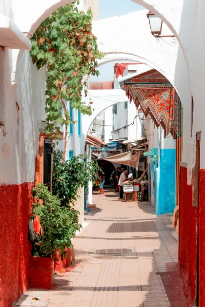 Scorcio di un vicolo stretto e soleggiato, incorniciato da archi bianchi, con pareti dipinte di rosso e blu, piante verdi e persone presso un mercato in lontananza.
