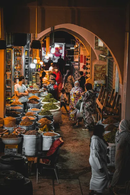 Un vicolo di mercato affollato di notte, dove le persone passano accanto a bancarelle piene di sacchi di spezie ed erbe aromatiche sotto archi illuminati.