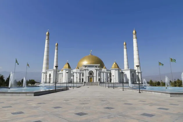 Una grande moschea bianca con una cupola dorata e quattro minareti si trova dietro una piazza pavimentata con fontane sotto un cielo azzurro e limpido.