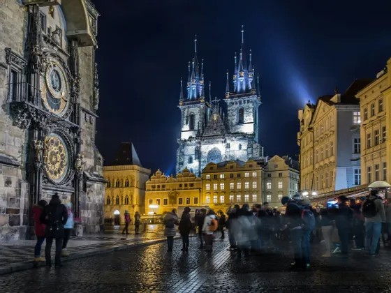 Una multitud de gente se congrega en una plaza adoquinada por la noche, contemplando una torre del reloj iluminada y una iglesia gótica.