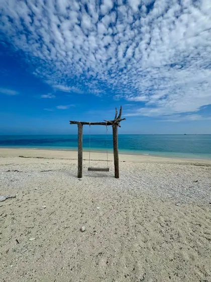 Un'altalena rustica in legno, vuota, su una spiaggia sabbiosa, di fronte all'oceano calmo e blu sotto un cielo parzialmente nuvoloso.