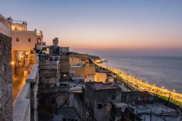 Una vista sui tetti di una storica città costiera al tramonto, con una strada illuminata che costeggia il mare.