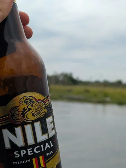 Un primo piano di una mano che tiene una bottiglia di birra Nile Special con un fiume e un paesaggio verde sullo sfondo.