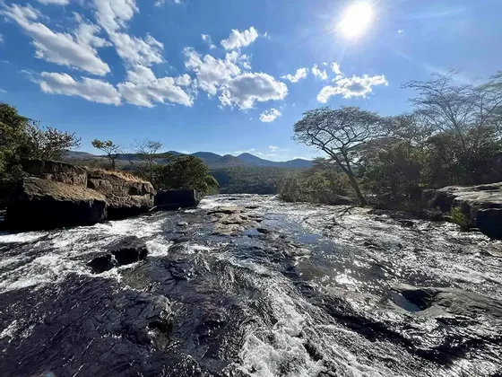 Un fiume ampio e roccioso scorre rapidamente sotto un sole splendente e un cielo azzurro nuvoloso, con colline boscose in lontananza.