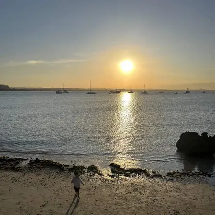 Un bambino piccolo cammina su una spiaggia sabbiosa al tramonto, con barche ancorate sull'acqua sullo sfondo.