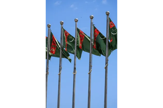 Una fila di bandiere del Turkmenistan che sventolano su aste d'argento contro un cielo azzurro chiaro.