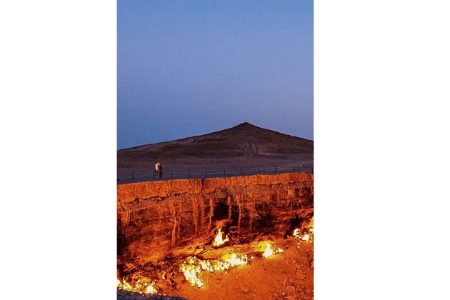 Due persone di un viaggio di gruppo WeRoad si trovano accanto a una ringhiera, guardando un grande cratere di gas infuocato nel deserto al tramonto.