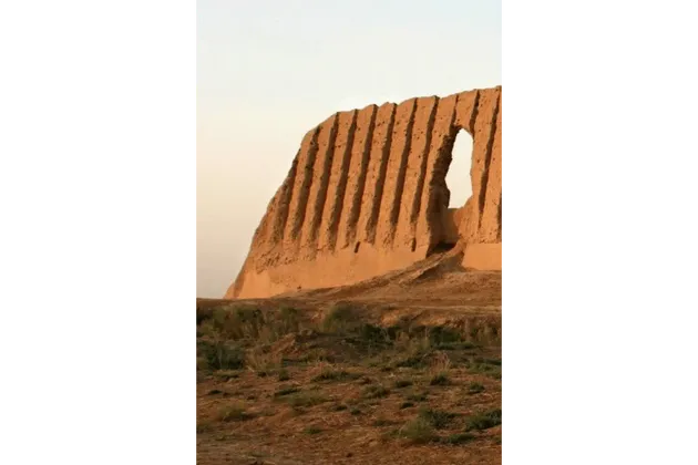 Una rovina di muro in mattoni di fango ondulato con una grande apertura si erge su una collina secca ed erbosa contro un cielo pallido e velato.
