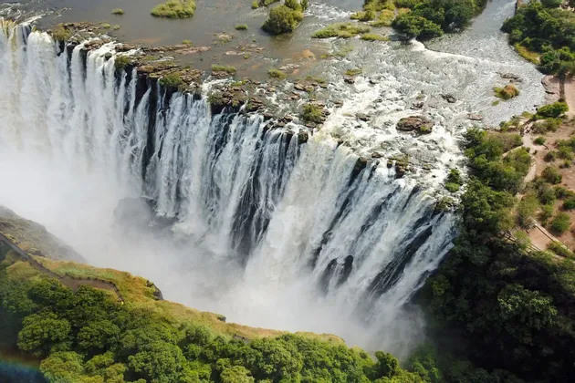 Una vista aerea di un'ampia cascata che si riversa da una scogliera rocciosa in una gola nebbiosa, circondata da alberi verdi.
