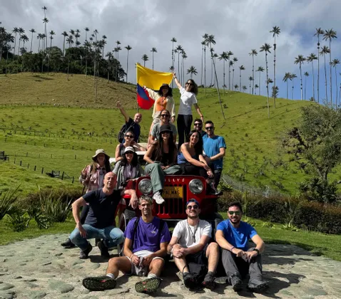 Un gruppo WeRoad in viaggio posa su e intorno a una jeep rossa in un paesaggio collinare con alte palme, con una persona che tiene una bandiera.