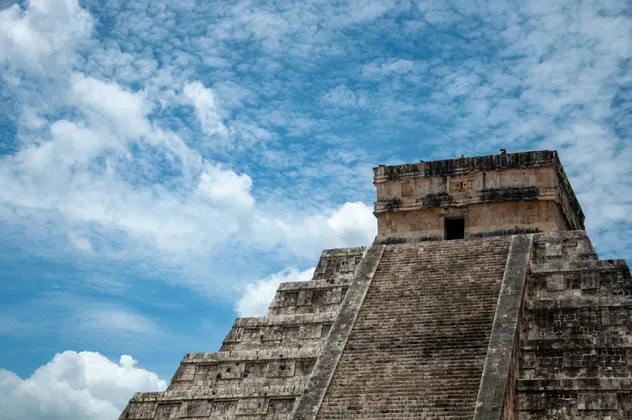 Una vista dal basso di un'antica piramide a gradoni in pietra che si eleva verso un cielo blu con nuvole bianche sparse.