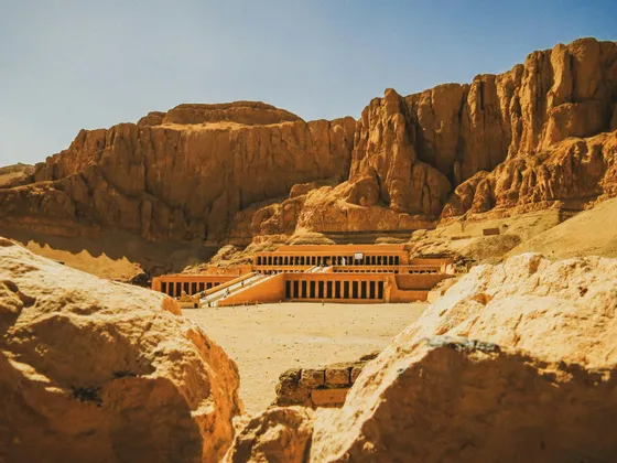 Un grande tempio terrazzato costruito alla base di un'imponente scogliera rocciosa, visto da tra due grandi massi sotto un cielo limpido.