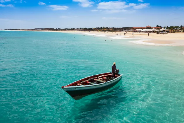Une personne portant un chapeau se tient dans une petite embarcation flottant sur une eau turquoise et limpide, près d'une plage de sable avec des gens.