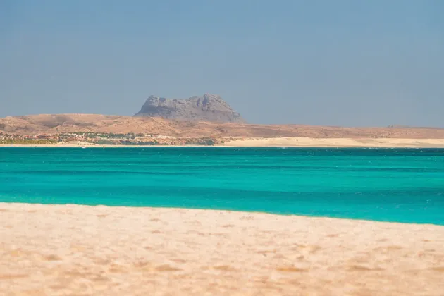 Une vue paysagère depuis une plage de sable, regardant par-dessus une eau turquoise vers une côte aride lointaine avec une montagne rocheuse.