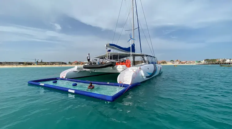 Un grand catamaran blanc ancré dans des eaux turquoise avec une piscine gonflable attachée, où une personne nage près d'une plage de sable.