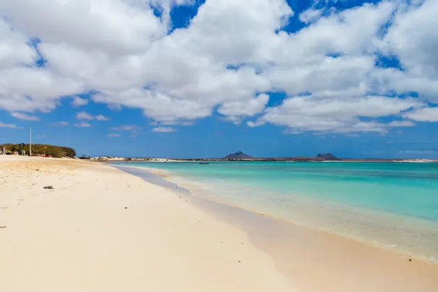 Une large plage de sable blanc rencontre une eau turquoise limpide sous un ciel bleu parsemé de nuages blancs.