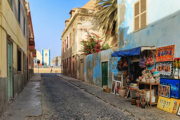 Une rue pavée tranquille bordée de bâtiments colorés et patinés, et un étal de marché vendant de l'artisanat sous un ciel clair.