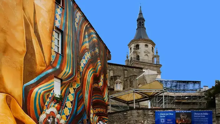 Un edificio con un grande murale colorato si erge accanto a un'antica torre campanaria in pietra sotto un cielo azzurro e limpido.