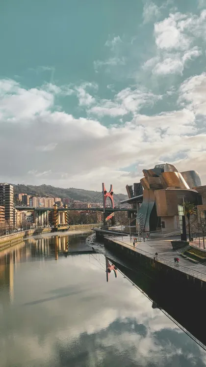 Un moderno edificio architettonico su una riva di fiume con il suo riflesso nell'acqua, un ponte rosso e una città sullo sfondo.