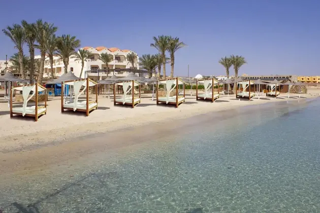 Lettini a baldacchino fiancheggiano una spiaggia sabbiosa con acqua cristallina di fronte a un resort alberghiero con palme.