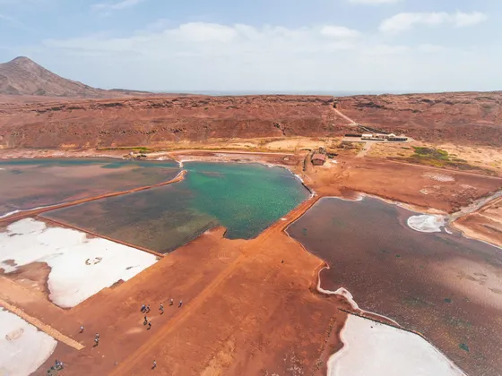 Une vue aérienne d'un voyage de groupe WeRoad marchant sur de la terre rouge le long de marais salants multicolores avec la mer en arrière-plan.