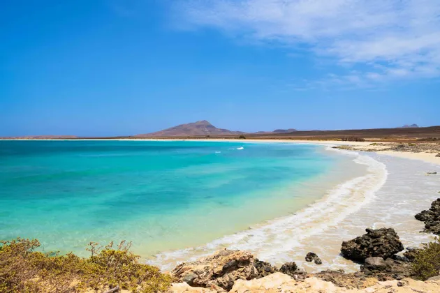 L'eau turquoise de l'océan lèche une plage de sable blanc avec des montagnes arides lointaines sous un ciel bleu clair.