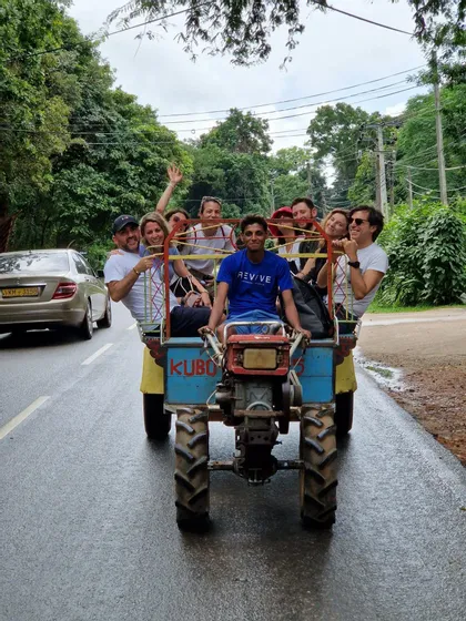 Eine WeRoad-Reisegruppe lächelt bei der Fahrt auf einem kleinen blauen Traktor auf einer nassen, von Bäumen gesäumten Straße.