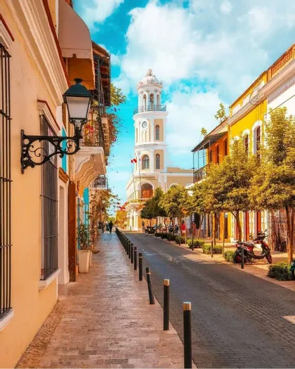 Una strada acciottolata illuminata dal sole, fiancheggiata da edifici colorati e alberi, che conduce verso un'imponente torre dell'orologio bianca sotto un cielo azzurro.
