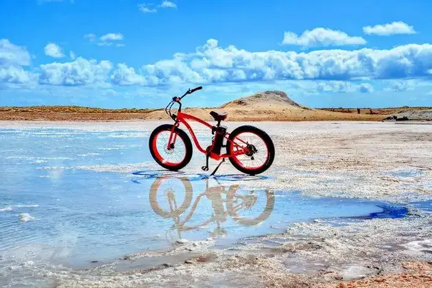Una e-bike rossa con gomme larghe è parcheggiata in acqua bassa su una salina, creando un chiaro riflesso sotto un cielo blu con nuvole bianche.