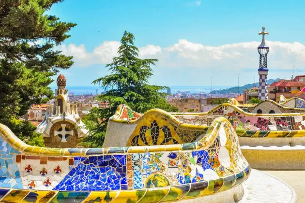 Una panca serpentina ricoperta di mosaici colorati si affaccia su uno skyline cittadino e sul mare, circondata da alberi verdi.