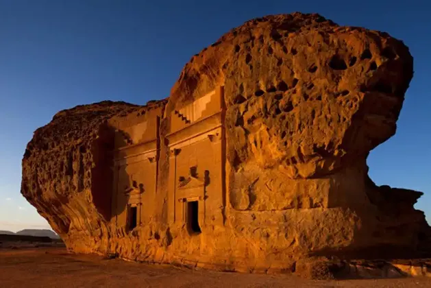Una facciata di antica tomba scavata in una grande roccia arenaria, illuminata da una calda luce solare sotto un cielo azzurro e limpido.