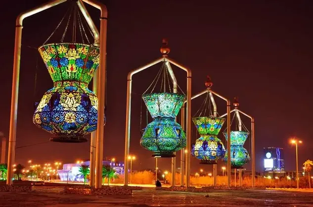 Una fila di grandi lanterne a mosaico colorate, appese a strutture e illuminate di notte, con lo skyline della città sullo sfondo.