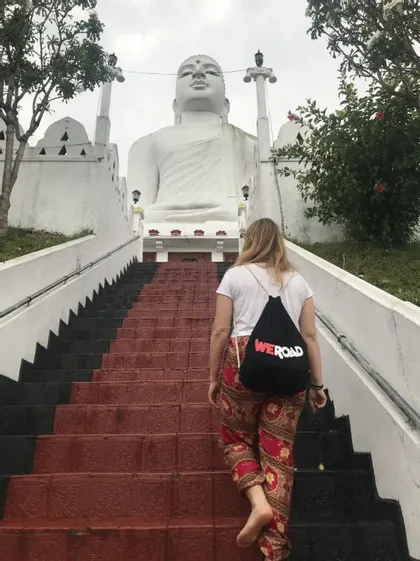 Una persona con uno zaino WeRoad cammina a piedi nudi su per una lunga scalinata verso una grande statua bianca di Buddha seduto.