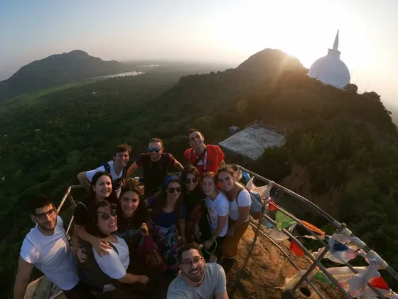 Un gruppo WeRoad scatta un selfie su una cima di montagna all'alba, con uno stupa bianco e una valle lussureggiante sullo sfondo.