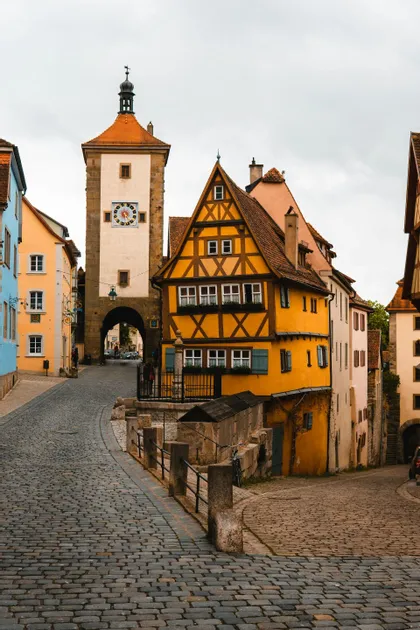 Una via acciottolata e tortuosa costeggia edifici colorati a graticcio e una torre dell'orologio in pietra in un centro storico.
