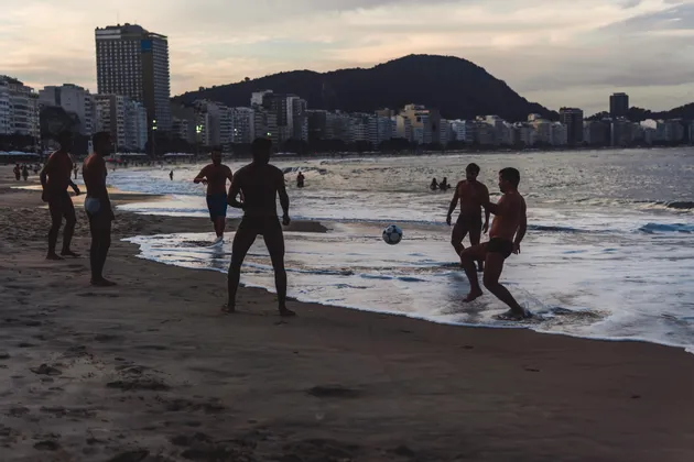 Un gruppo di uomini in silhouette che giocano a calcio sulla riva di una spiaggia al tramonto, con lo skyline di una città sullo sfondo.