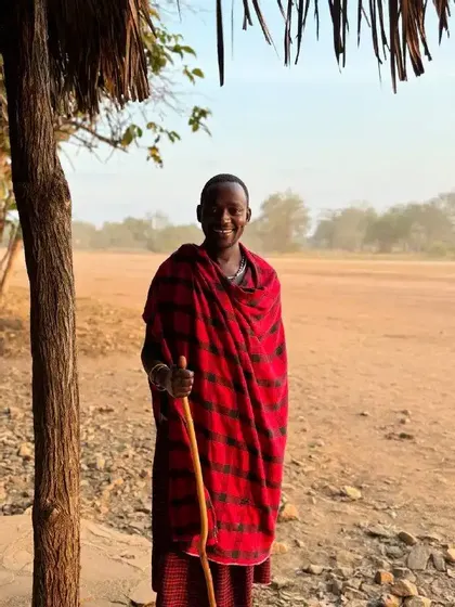 Un uomo sorridente con una tunica scozzese rossa e nera tiene un bastone da passeggio mentre è in piedi accanto a un albero in un paesaggio polveroso.