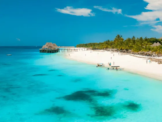 Vista aerea di una spiaggia di sabbia bianca con acqua turchese, palme e un pontile di legno con una palafitta sotto un cielo blu.