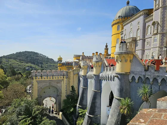 Un palais coloré avec des tours jaunes et grises, des gens se promenant le long de ses remparts sous un ciel bleu à côté d'une colline verdoyante.