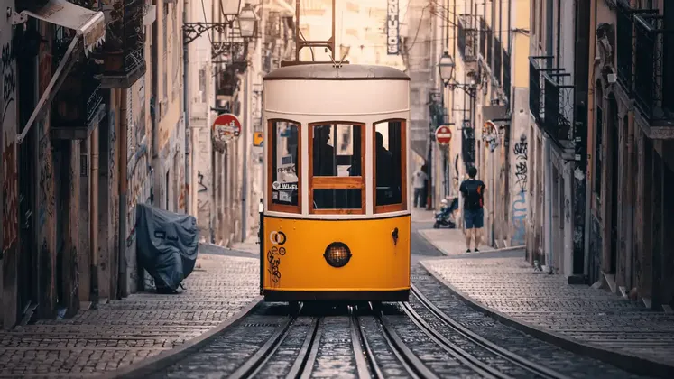 Un tramway vintage jaune et blanc roule sur des rails dans une rue étroite pavée bordée de vieux bâtiments.