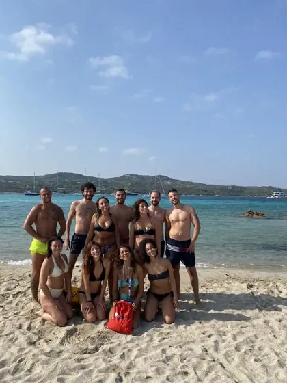 Un viaggio di gruppo WeRoad con uomini e donne in costume da bagno in posa per una foto su una spiaggia sabbiosa, con acqua turchese e barche a vela sullo sfondo.