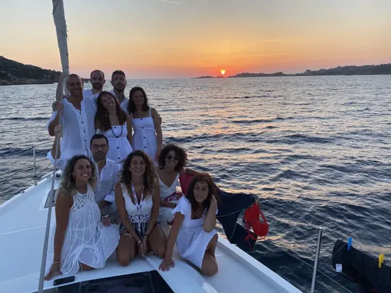 Un viaggio di gruppo WeRoad, tutti vestiti di bianco, in posa insieme sul ponte di una barca a vela al tramonto.