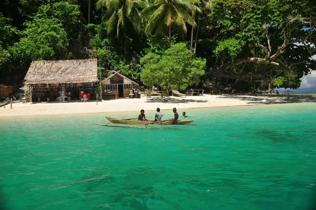 Tre persone remano una piccola canoa in acqua turchese al largo di una spiaggia di sabbia bianca con capanne di paglia e una giungla fitta.