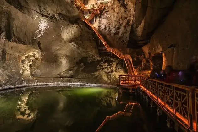 A large, illuminated cavern with a wooden staircase climbing the rock wall and a walkway next to a dark pool of water.