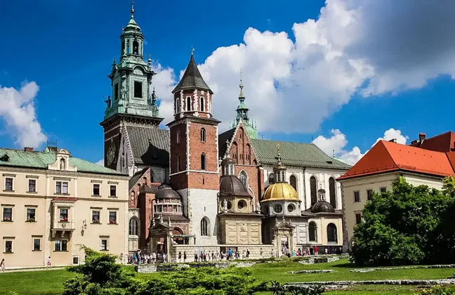 A historic cathedral complex with brick towers, green spires, and a golden dome sits on a grassy hill under a blue sky with clouds.